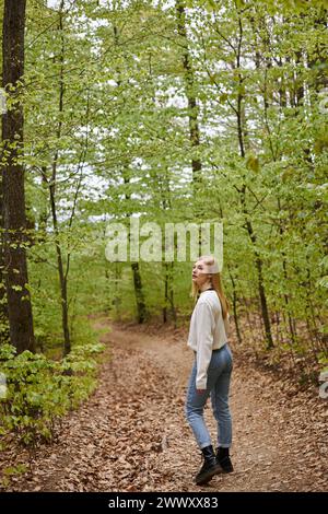 Back view image of curious blonde female traveler hiking in forest in sweater and jeans Stock Photo