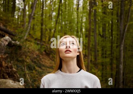 Relaxed blonde woman breathing fresh air in forest on solo hiking trip looking in the sky Stock Photo