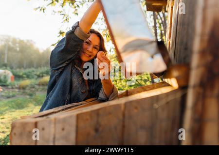 Happy middle-aged woman in hat collects fresh egg from a chicken coop at dawn. Stock Photo