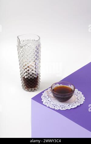a violet cube made with colored paper to create a 3D optical illusion. On top is a vintage pink coffee cup with a white lace doilyand a glass jug of c Stock Photo