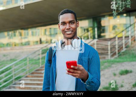 Individual portrait of one african american guy looking at camera and holding a smartphone. Real teenage high school student using a cellphone and staring front at university campus. Man with phone. High quality photo Stock Photo
