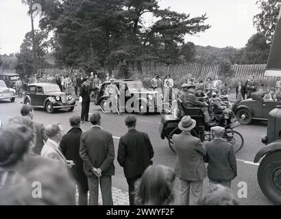 1950s, historical, spectators roadside watching a veteran car drive off on a vintage car run, with a British policeman standing in the middle of the road signaling the other road users to wait, England, UK. Stock Photo