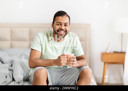 Cheerful african american man with coffee sitting on bed Stock Photo