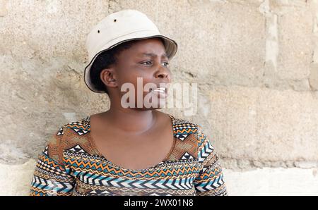 young african woman standing next to a wall she is wearing a hat, village life Stock Photo