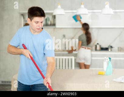 Young man in home clothes makes kitchen floor perfectly clean using mop Stock Photo