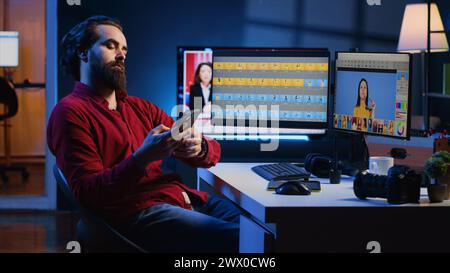 Photographer uses retouching software to edit pictures on computer monitors, taking a break after working too much. Photo editor tired after doing color grading on images, relaxing by using smartphone Stock Photo