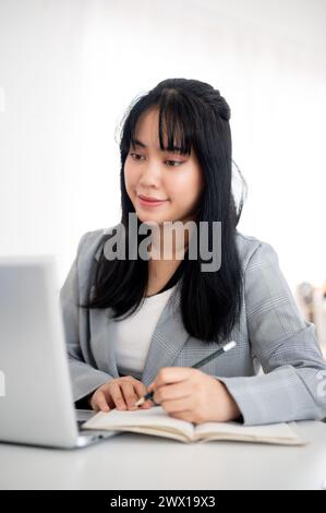A young Asian businesswoman or female office worker is looking at her laptop screen while taking notes in the notebook, working in a modern office. Stock Photo