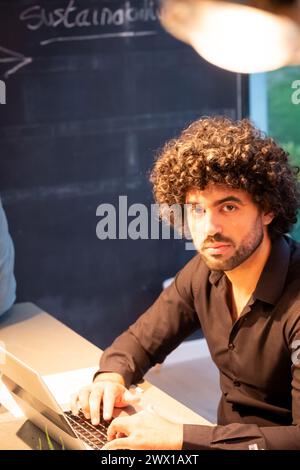 A young professional man, possibly of Middle Eastern or Hispanic descent, is captured in a moment of concentration at his workplace. With curly hair and a black shirt, he appears engrossed in his work on a laptop, reflecting determination and dedication. The background features a chalkboard with the word Sustainability written on it, hinting at the nature of his work or the values of the organization. The warm overhead lighting accentuates his features and the intent focus in his eyes. Focused Professional at Work. High quality photo Stock Photo