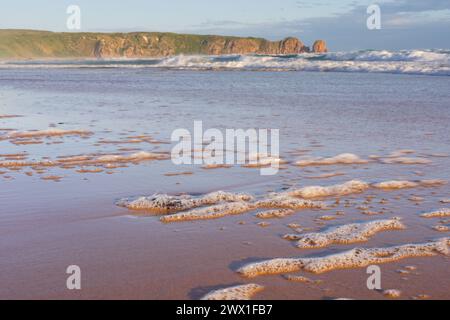 Low angle view along a beach covered in frothy bubbles with a rocky headland in the distance at Cape Woolamai on Phillip Island in Victoria, Australia. Stock Photo