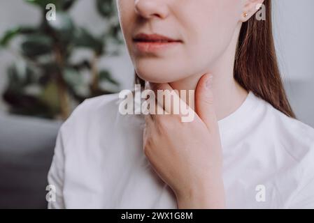 Close up of sad young woman with sore throat sitting alone on couch, pain thyroid gland on neck or disease reflux, acid of suffer people. Sickness in Stock Photo