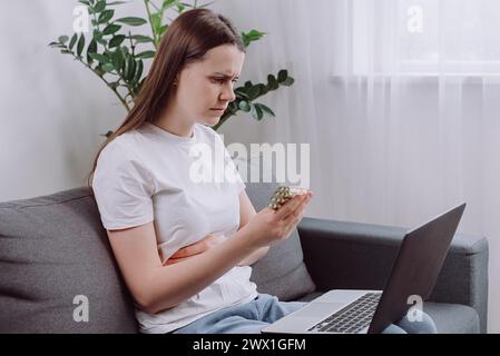 Wellness and dieting concept. Sad young brunette woman using computer sitting alone on sofa at home, holding pills tablet pack read prescription medic Stock Photo