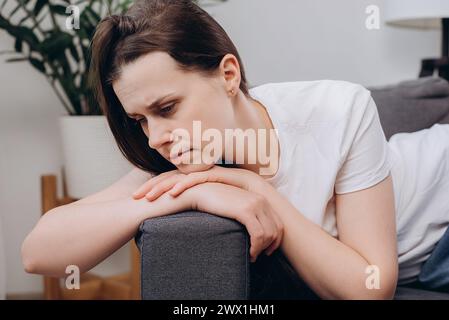 Close up of pensive stressed young caucasian woman sitting alone on sofa at home, looking away, thinking over problems, concern, touching face, feelin Stock Photo