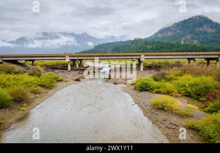 A braided shallow stream runs beneath a concrete highway bridge with yellow autumn shrubs along its banks and low cloud over hills in Washington. Stock Photo