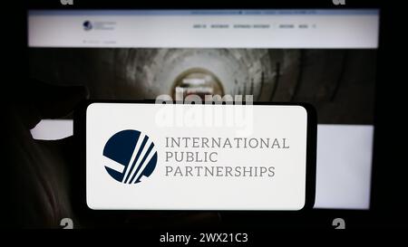 Person holding cellphone with logo of British company International Public Partnerships Limited in front of webpage. Focus on phone display. Stock Photo