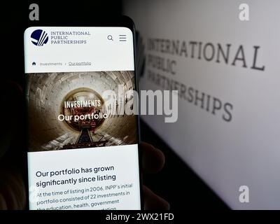 Person holding cellphone with webpage of British company International Public Partnerships Limited with logo. Focus on center of phone display. Stock Photo