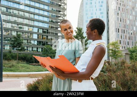 Two business colleagues talk while holding a folder between their hands, situated in a park in a business district area. Stock Photo