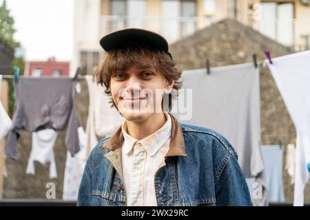 Smiling young man in denim jacket standing in front of hanging laundry. Stock Photo
