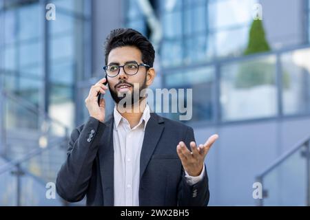 Close-up photo of a busy young Indian businessman man in a suit and glasses standing near an office center on the street, talking seriously on the phone and gesturing with his hand. Stock Photo