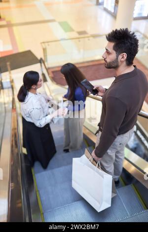 Vertical side view of bearded adult man holding mobile phone while standing on escalator in shopping mall Stock Photo