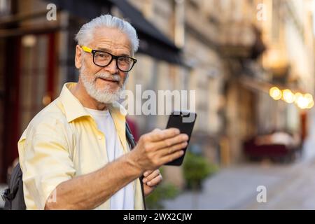 Portrait of a senior gray-haired smiling man walking and traveling in the city with a rucksack, holding a phone in his hand and looking happily at the camera. Stock Photo