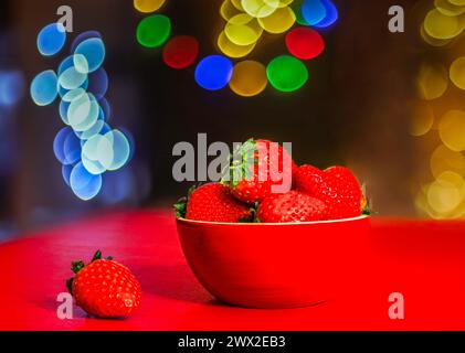 Heap of fresh strawberries in a red ceramic bowl on a red table and colorful bokeh Stock Photo