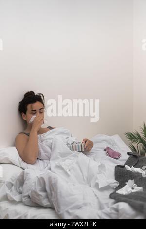 In bed, an ill young woman wipes her nose with tissues, holding medical drops to ease her illness Stock Photo