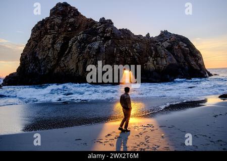 Sunset illuminates person in front of Window in the Rock feature of Keyhole Arch at Pfeiffer Beach in Big Sur on the Pacific Ocean in California, USA. Stock Photo