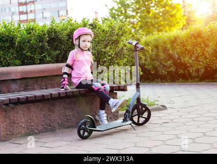 A beautiful seven-year-old girl sits on a bench in pink protective equipment next to a scooter. The concept of cycling and scootering without injury. Stock Photo
