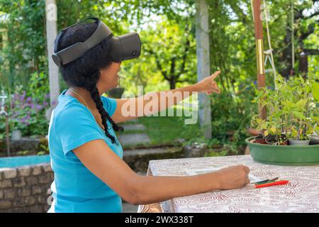 A multiracial woman, wearing virtual reality glasses, sits at a garden table, pointing and creating a garden plan. Stock Photo