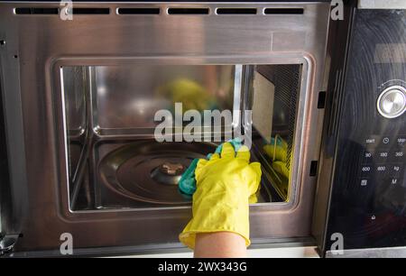 A girl's hand in a yellow glove with a sponge washes the microwave oven from grease and dirt. Foam and anti-grease. Stock Photo