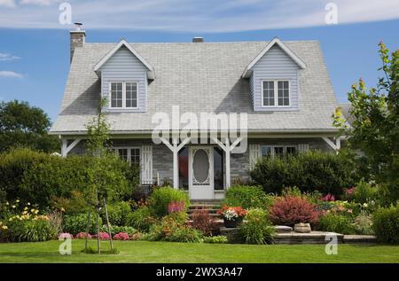 Facade of old grey nuanced brick with white trim cottage style Canadiana home with dormer windows and landscaped front yard with mixed perennial Stock Photo