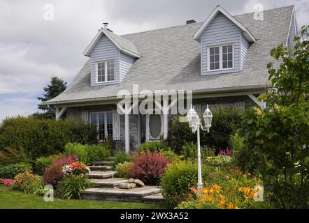 Facade of old grey nuanced brick with white trim cottage style Canadiana home with dormer windows and landscaped front yard with mixed perennial Stock Photo