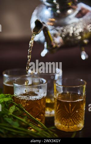 Close-up of pouring delicious Moroccan mint tea from a silver teapot into drinking glasses. Bunch of fresh mint in the foreground. Tea break in Morocc Stock Photo