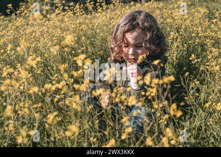 Happy and cheerful little boy playing with yellow flowers, sitting on a green pad full of petals on bright sunny day Stock Photo