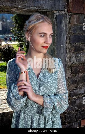 On a sunny day, a young woman with blonde hair stands in the middle of an idyllic landscape. She is wearing an airy dress that emphasises her delicate Stock Photo