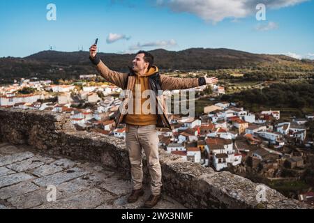 young happy photographer man traveling sightseeing in a small town, taking a selfie of the panoramic view of the town and the mountains, from a castle Stock Photo