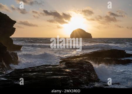 The sun setting behind Gull Rock viewed from Trebarwith Strand Beach, North Cornwall UK. Evening scene with waves, surf and jagged cliffs. Stock Photo