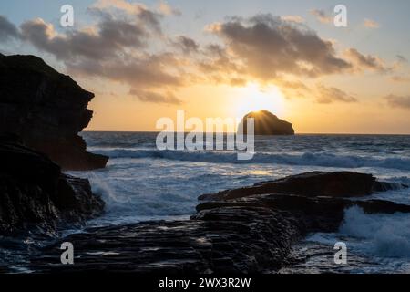 The sun setting behind Gull Rock viewed from Trebarwith Strand Beach, North Cornwall UK. Evening scene with waves, surf and jagged cliffs. Stock Photo