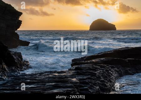 The sun setting behind Gull Rock viewed from Trebarwith Strand Beach, North Cornwall UK. Evening scene with waves, surf and jagged cliffs. Stock Photo