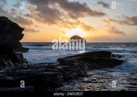 The sun setting behind Gull Rock viewed from Trebarwith Strand Beach, North Cornwall UK. Evening scene with waves, surf and jagged cliffs. Stock Photo