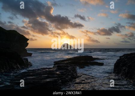 The sun setting behind Gull Rock viewed from Trebarwith Strand Beach, North Cornwall UK. Evening scene with waves, surf and jagged cliffs. Stock Photo