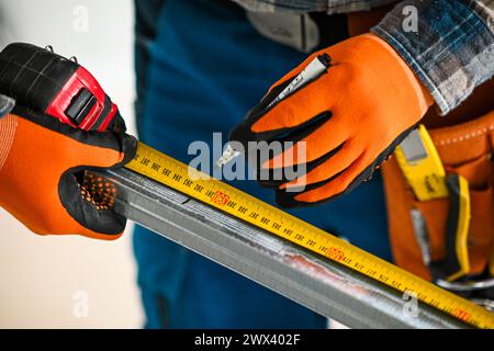 a construction worker in overalls, gloves and a tool belt measures the length of a plasterboard profile with a meter - close-up view. Stock Photo