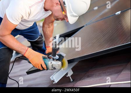 Worker installing or maintaining photovoltaic solar panel system on roof of house. Close up technician using grinder on metal part of the panel's mounting structure. Concept renewable energy. Stock Photo