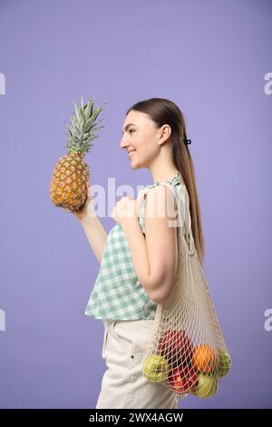 Woman with string bag of fresh fruits holding pineapple on violet background Stock Photo