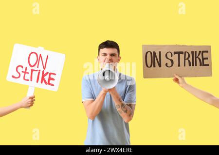 Protesting young man shouting into megaphone and placards with text ON STRIKE against yellow background Stock Photo