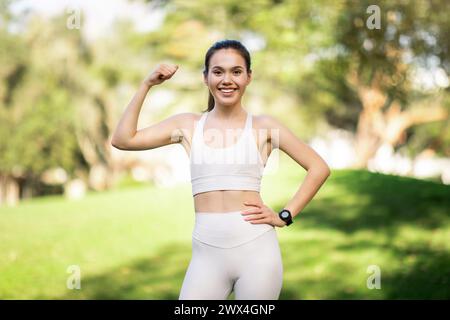 A radiant young woman in a white sports bra and leggings flexes her arm Stock Photo