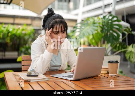 A stressed, serious Asian woman is working remotely at an outdoor space, focusing on reading an online document on her laptop. Stock Photo