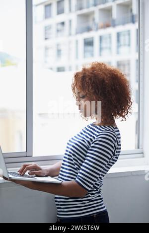 Office, window and black woman typing on laptop with research or online communication on email. Creative, employee and working with computer to scroll Stock Photo