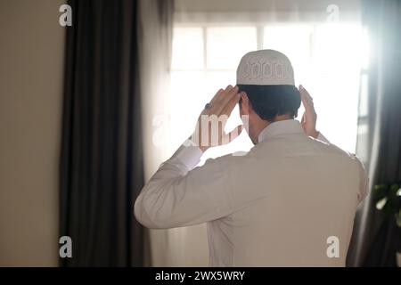 Rear view of unrecognizable young Muslim man wearing white thobe and taqiyah praying salah, copy space Stock Photo
