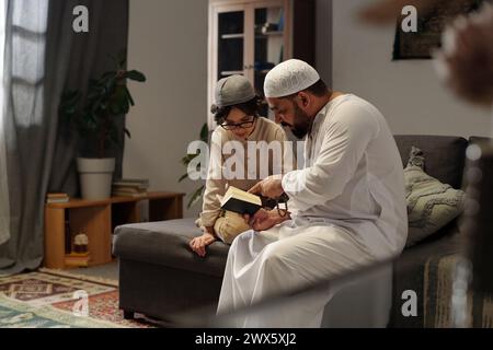 Modern bearded Muslim man wearing kandora sitting on sofa in living room explaining Quran surah to his son, copy space Stock Photo